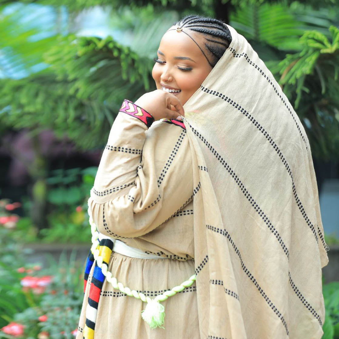 Woman in traditional attire with a headscarf, standing outdoors with greenery in the background, ethiopian clothing